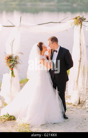 Schöne Braut im weißen Kleid und schönen Bräutigam trägt schwarzen Anzug umarmt unter Torbogen am Strand-Hochzeits-Zeremonie in der Nähe von See Stockfoto