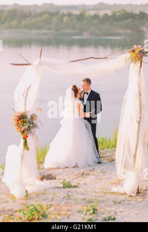 Schöne Braut im weißen Kleid und schönen Bräutigam trägt schwarzen Anzug umarmt unter Torbogen am Strand-Hochzeits-Zeremonie in der Nähe von See Stockfoto