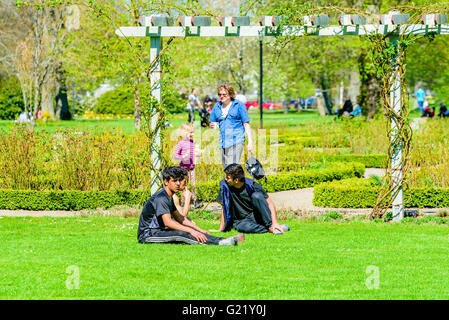 Ronneby, Schweden - 8. Mai 2016: Drei junge Erwachsene Männer sitzen auf der Wiese im öffentlichen Park die Sonne zu genießen. Frau und Kind Stockfoto