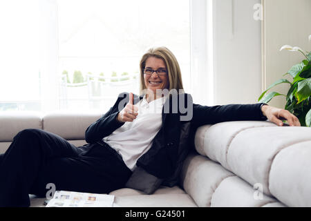 Porträt einer attraktiven Geschäftsfrau mittleren Alters, die zu Hause auf dem Sofa sitzt und für die Kamera lächelt und die Daumen hoch hält. Stockfoto