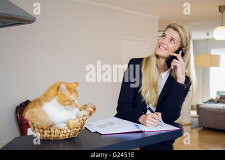 Attraktive business Frau mittleren Alters mit einem Telefon- und Notizen zu machen. Rothaarige Katze in geflochtenen Korb neben ihr sitzt. Entspannen Stockfoto