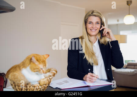 Eine Frau mittleren Alters in Anzug gerne Gespräche am Telefon, während Ihr Haustier Katze auf dem Zähler neben ihr sitzt in einem Weidenkorb. Stockfoto