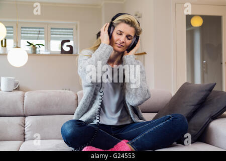 Blonde Frau mittleren Alters das Hören von Musik über Kopfhörer, entspannt zu Hause. Mittellange Aufnahme. Stockfoto