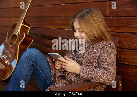 blonde junge Mädchen spielen Smartphone und Gitarre auf hölzernen Hintergrund Stockfoto