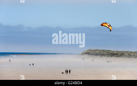 Menschen zu Fuß und einem Drachen genießen Sie das Wetter am Strand von Bamburgh, Northumberland als Meer Bund Rollen in. Stockfoto