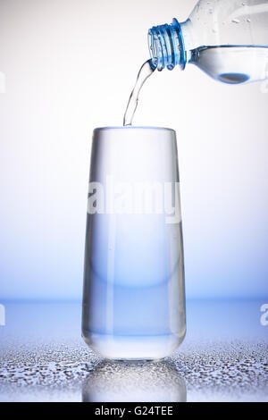Wasser gießen aus Kunststoff-Flasche in voller Trinkglas mit Spiegelung auf Tropfen des Wassers auf weißen und blauen Hintergrund Stockfoto