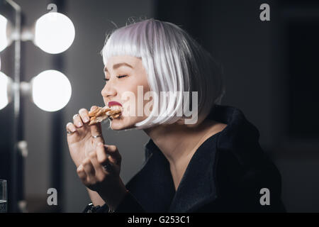 Lustige nette junge Frau in blonde Perücke Essen Pizza in Umkleidekabine Stockfoto