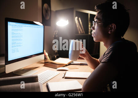 Profil von hübschen asiatischen jungen Mann studieren mit Computer und Teetrinken im dunklen Raum Stockfoto