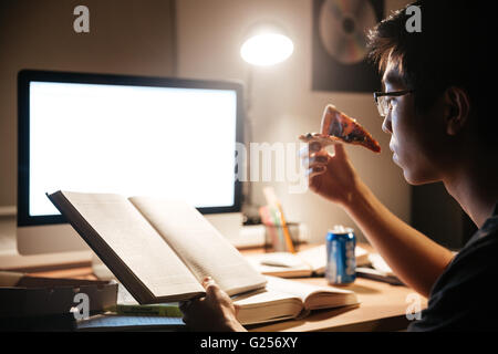 Konzentrierte sich asiatische jungen Mann in Gläsern Pizza essen und nachts zu Hause lesen Stockfoto