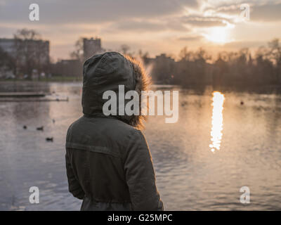 Eine Person trägt einen dicken warmen Mantel steht an einem Teich in einem Park und den Sonnenuntergang im winter Stockfoto