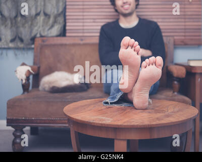 Barfuß junger Mann ruht seine Beine auf einem Kaffeetisch zu Hause, es ist eine Katze auf dem Sofa neben ihm Stockfoto