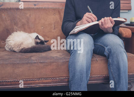 Ein junger Mann sitzt auf einem Sofa mit einer Katze und ist in ein Notizbuch schreiben Stockfoto