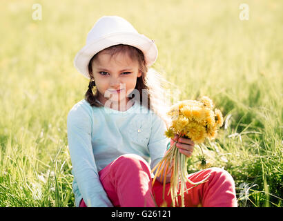 Grimassierende Kind Mädchen in Mode Hut sitzen auf dem Rasen und gelbe Blüten halten Stockfoto
