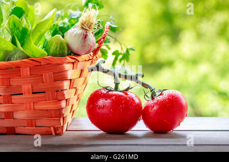 Geflochtenen Korb voller frisches Gemüse auf Holzoberfläche mit Tomaten im Fokus daneben. Grüner Hintergrund mit Textfreiraum Stockfoto