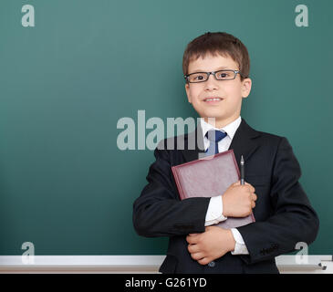 verwirrt Schuljunge mit Buch Portrait, gekleidet in klassischen schwarzen Anzug, auf grüne Tafel Hintergrund, Bildung und Wirtschaft co Stockfoto