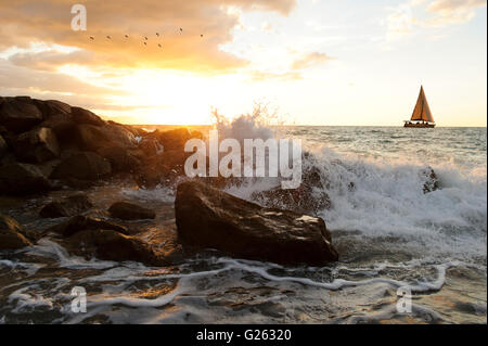 Segeln ist ein Segelboot segeln am Meer entlang mit einer Welle, die an den Ozean Felsen wie ein Schwarm Vögel fliegen in den Sonnenuntergang Himmel brechen. Stockfoto