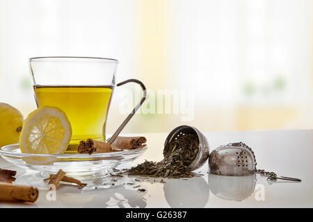 Glas Tasse Grüntee mit Zimtstange und Zitrone auf einem Tisch im Wohnzimmer. Mit metallischen Sieb horizontale Komposition. Stockfoto