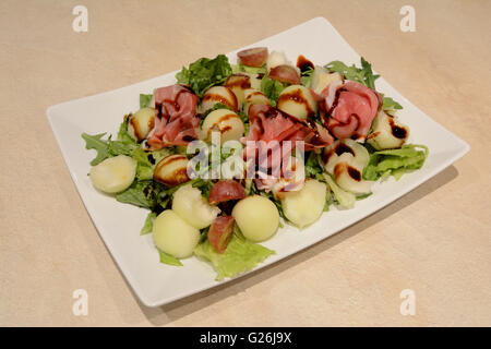 Quadratische Platte mit Salat Kuhmist, Melone, Trauben, Schinken und Balsamico-Essig Stockfoto