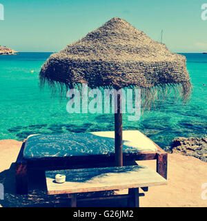 Detail der Entspannungsbereich an einem Strand in Ibiza, Spanien, mit einer bequemen Sonnenliege und eine rustikale Sonnenschirm hergestellt aus natürlichen Fasern Stockfoto