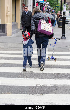 Eine Frau kreuzt die Straße an einem Fußgängerüberweg mit einem kleinen Kind. Sie trägt das Kind Roller. Stockfoto