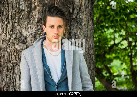 Hübscher junger Mann lehnte sich gegen Baum, Blick in die Kamera, an einem sonnigen Tag trägt einen leichten grauen Mantel Stockfoto