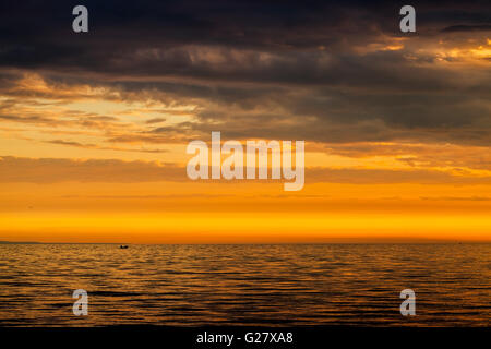 Dramatische Wolkengebilde, helle Sonnenuntergang über der Ostsee. Bewölkter Himmel, stilles Wasser und Fischer auf einem kleinen Boot in der Ferne Stockfoto