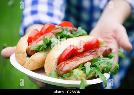 Frauenhand holding gegrillt Wurstbrote mit Rucola und Tomaten auf einem Teller Whitepaper auf Rasen Hintergrund Stockfoto