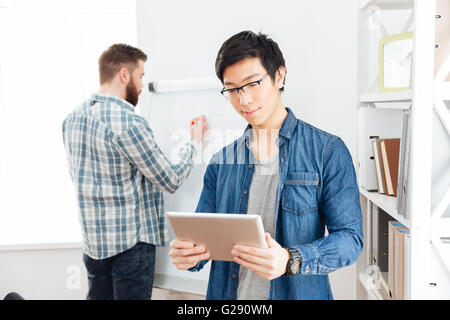 Schweren asiatischen Jüngling in Gläsern mit Tablet während sein Kollege auf Flipchart im Büro schreiben Stockfoto