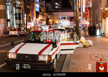 Japan, Kumamoto Stadtzentrum. Abend. Taxi-Cab durch Pflaster mit offener Tür in den Vordergrund. Blick entlang der Straße, Verkehr hinter dem Taxi, einige Leute. Stockfoto