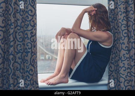 Eine junge Frau sitzt auf dem Fensterbrett und sucht sich an einem regnerischen Tag Stockfoto