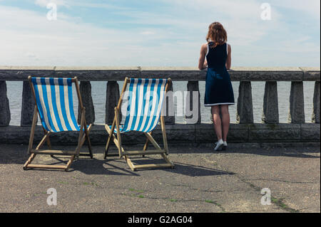 Eine junge Frau steht von einer Mauer mit Beton Balustraden an einer Promenade und das Meer bewundern ist, gibt es zwei Liegestühle Stockfoto