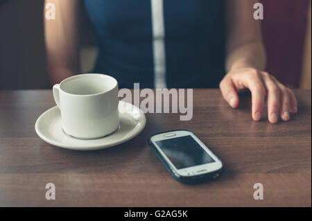 Eine junge Frau sitzt am Fenster in einem Diner mit einer Tasse und einem Smartphone vor ihr Stockfoto