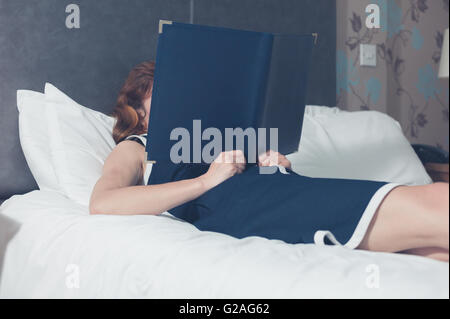Eine junge Frau liegt auf einem Bett in einem Hotelzimmer und liest die Hotelinformationen Stockfoto