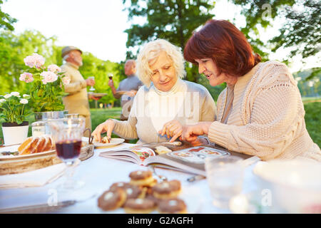 Zwei ältere Frauen lesen Kochbuch beim Essen Kuchen in einem Garten Stockfoto