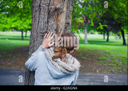 Eine junge Frau verbirgt sich hinter einem Baum im park Stockfoto