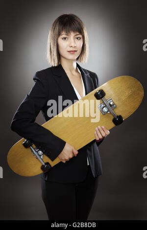 professionell aussehende Geschäftsfrau hält eine Skateboard gedreht im Studio auf einem grauen Hintergrund niedrig wichtige Beleuchtung Stockfoto