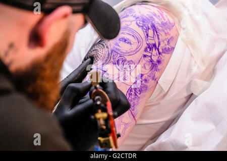 BELFAST, NORDIRLAND. 28. MAI 2016 - Nordirische Tattoo-Konferenz 6. Ein Tätowierer zeichnet ein kompliziertes Tattoo einer Frau, die eine Gasmaske am Arm einer Kundin trägt Stockfoto