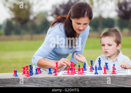 Elegante junge im weißen Hemd und seine Mutter, mit blauen und roten Schachfiguren Schach auf Holztisch im Park spielen lernen Stockfoto