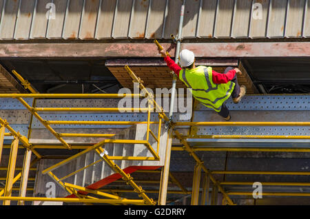 Ein weißer Arbeiter in gut sichtbarer Kleidung (Hi-vis), der auf dem Stahldach einer mehrstöckigen Baustelle auf einem gelb gefärbten Gerüst klettert Stockfoto