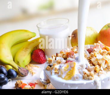 Strom von Milch in eine Schüssel Müsli und Früchte auf einem Tisch mit Früchten und Glas Milch Hintergrund in der Küche fallen. Stockfoto