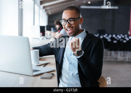 Hübscher junger Geschäftsmann sitzt an seinem Ddesk feiert Erfolg mit beim Betrachten seiner Laptop-Bildschirm erhobenen Armen Stockfoto