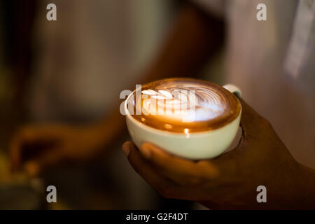 Gewusst wie: Latte Art Kaffee zu machen. Hände halten eine Tasse Kaffee mit Schaum über Holztisch, Seitenansicht Stockfoto