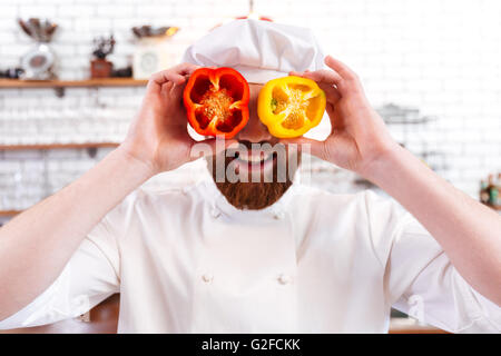 Spielerische bärtigen Chef-Koch holding Hälften des roten und gelben Paprika vor seinen Augen und Lächeln Stockfoto