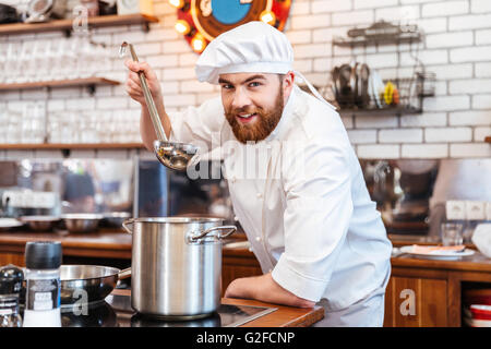 Fröhliche bärtigen Koch Kochen Kochen und schmecken Suppe aus der Pfanne mit Kelle auf die Küche Stockfoto