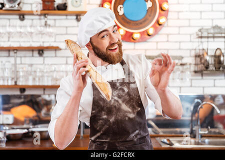 Glücklich schönen Bäcker Brot hält und ok Anzeichen auf die Küche Stockfoto