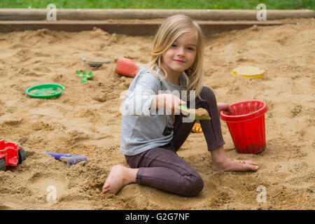 Kleines blondes Mädchen spielt im Sandkasten mit Spielzeug aus Plastik-tools Stockfoto