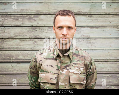 Ernsthafte kaukasischen Jüngling in militärischen Tarnuniform. Outdoor Closeup Portrait über grüne ländliche Holzwand Stockfoto
