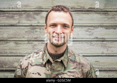 Lächelnde kaukasischen Jüngling in militärischen Tarnuniform. Outdoor Closeup Portrait über grüne ländliche Holzwand Stockfoto