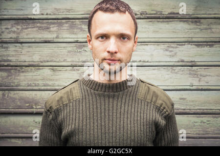 Schönen jungen kaukasischen Mann im militärischen grünen Pullover. Outdoor Closeup Portrait über grüne ländliche Holzwand Stockfoto