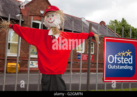 Strohpupille mit rotem Hemd und Mütze. Schuljungen Vogelscheuche vor der Schule mit Ofsted-Schild für hervorragende Inspektion, Lancashire, Großbritannien. Stockfoto